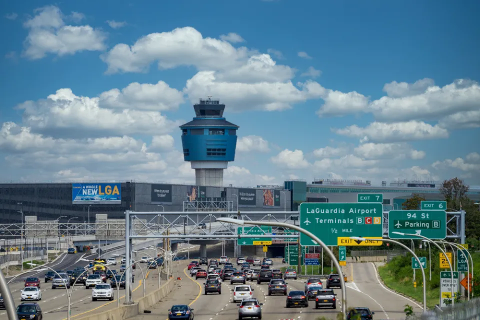 Freeway with traffic leading to LaGuardia Airport with air control tower in the background against white fluffy clouds on bue sky
